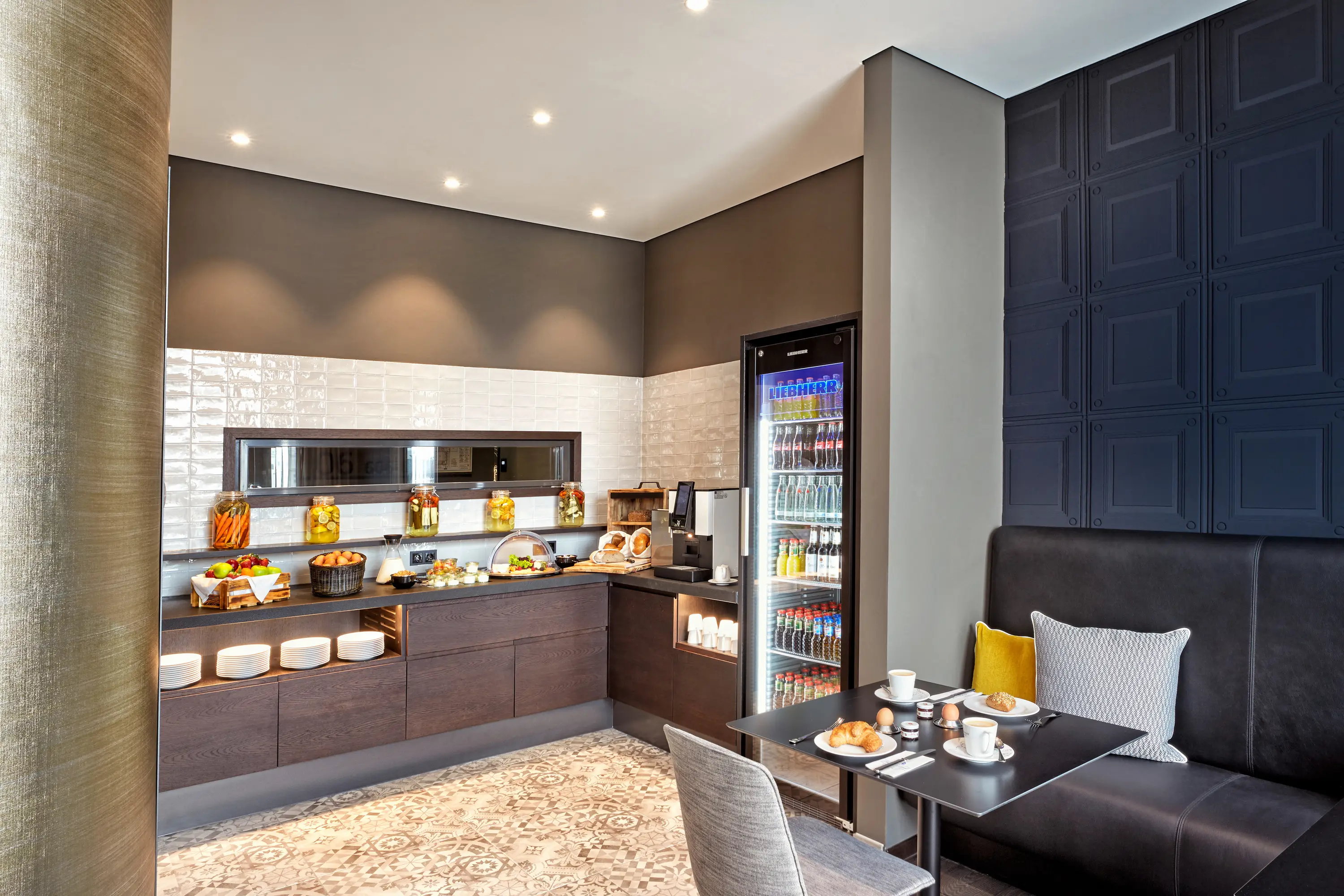 Küchenecke mit kleinen Snacks, einer Kaffeemaschine und einem großen Getränkekühlschrank. Daneben steht eine schwarze Ledercouch und ein Stuhl. Küchenecke mit kleinen Snacks, einer Kaffeemaschine und einem großen Getränkekühlschrank. Daneben steht eine schwarze Ledercouch und ein Stuhl.